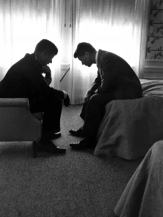 John and Robert Kennedy sitting face to face in silhouette against a bright hotel window, heads bowed