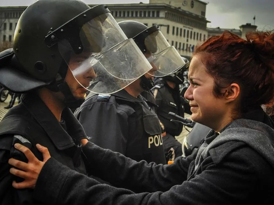 A crying young woman with red hair holding the shoulders of a riot policeman in a helmet and visor, in a police cordon in Sofia