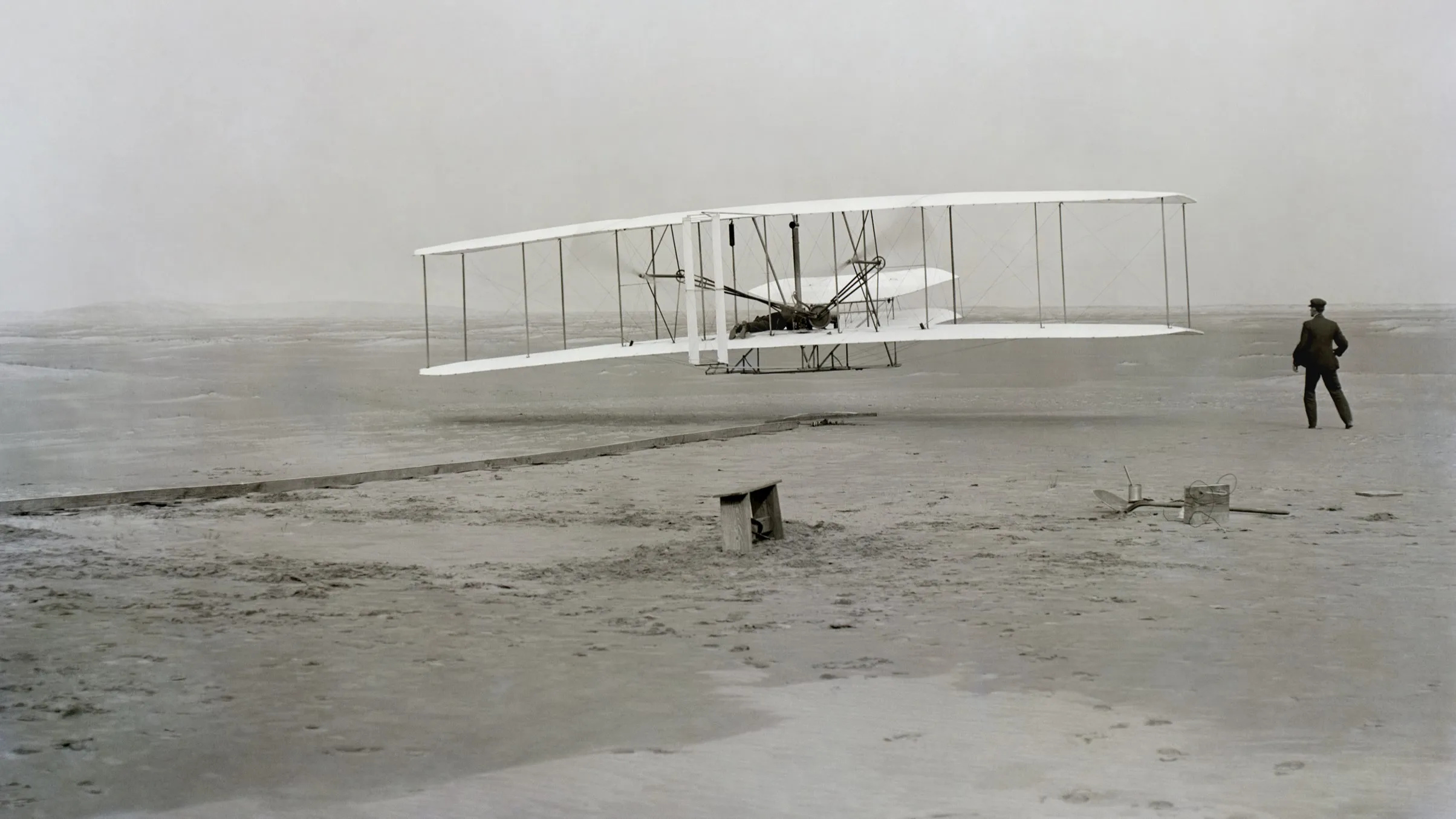 The Wright Flyer lifting off its wooden launching rail a few feet above the sand, Orville Wright lying at the controls and Wilbur Wright standing to the right