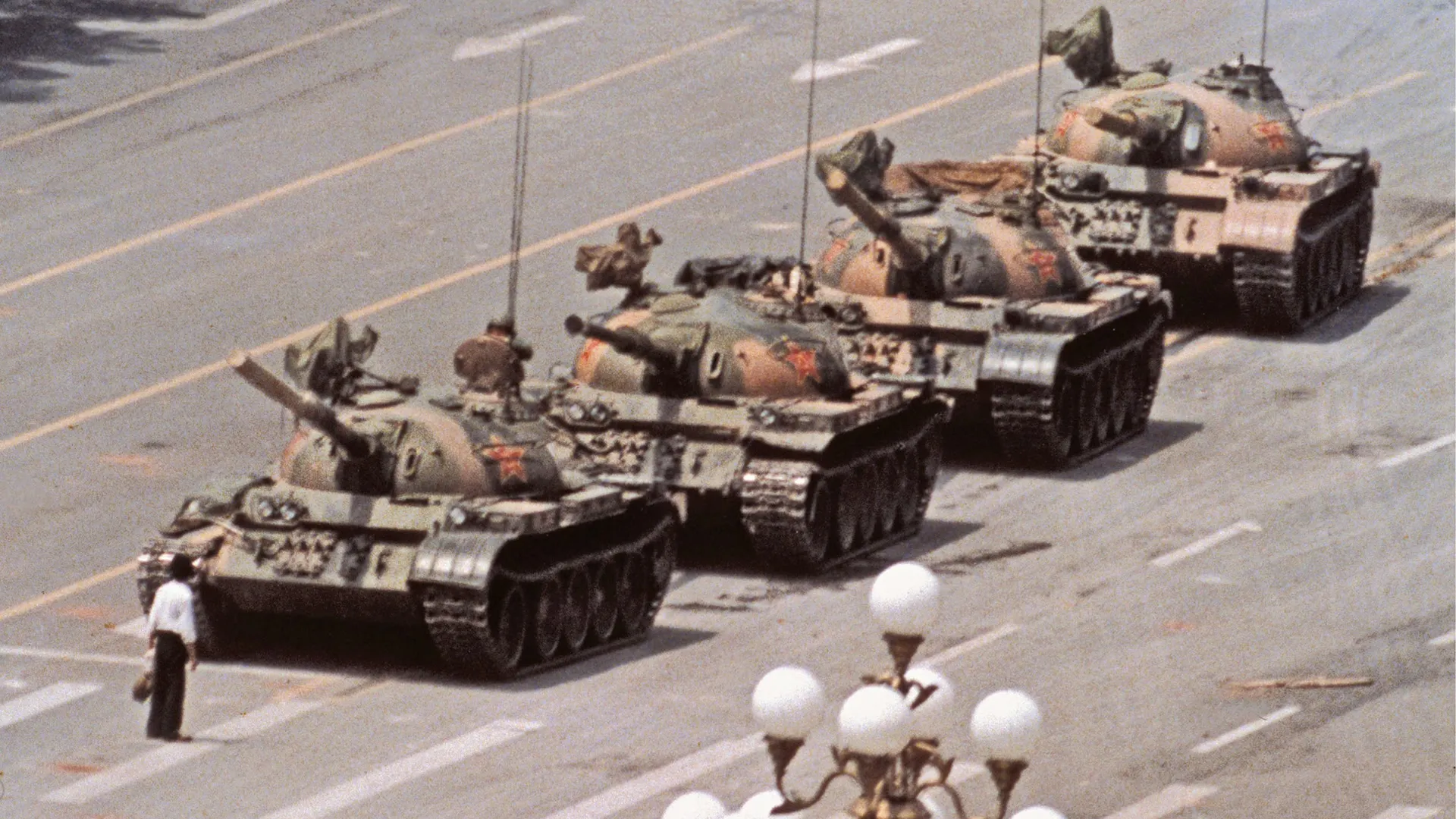 A lone man in a white shirt, a bag in his hand, standing in front of a column of four tanks on an empty avenue in Beijing